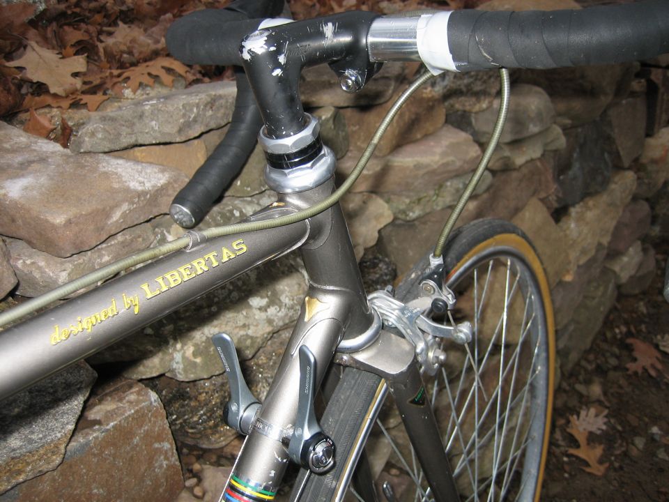 Silver road bike front detail with drop bars, quill stem, and 'designed by LIBERTAS' decal against a stone wall.