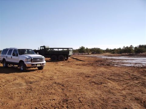 1978 Ford Bronco - A white Ford Excursion and a military truck are parked in a muddy, open field under a clear blue sky.