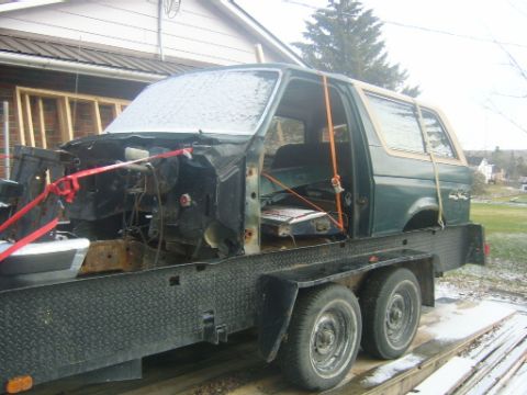 1990 Ford Bronco - The body of a green 1990 Ford Bronco is secured to a flatbed trailer, with its front end disassembled.