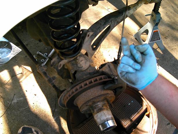 close-up of rusty vehicle suspension components, including a coil spring, brake rotor, and hub assembly, with a gloved hand pointing