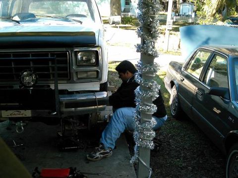 1986 Ford Bronco - A man works on the front suspension of a white Ford truck, with a dark sedan parked nearby.