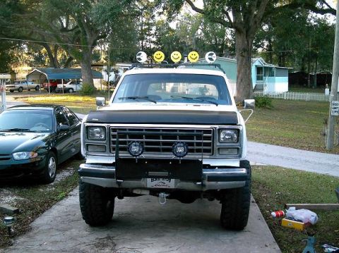 1986 Ford Bronco - A lifted white SUV with a black hood decal and decorative smiley face lights on its roof rack is parked in a driveway.