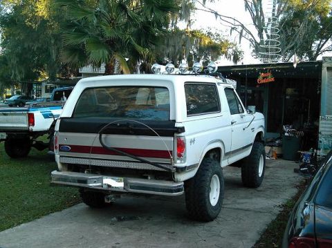 1986 Ford Bronco - A lifted white Ford Bronco sits parked next to a pickup truck, with a Christmas decoration visible in the background.