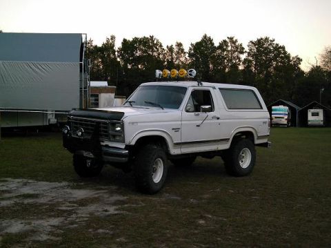 1986 Ford Bronco - A white Ford Bronco with a suspension lift and auxiliary lights sits in a grassy field.