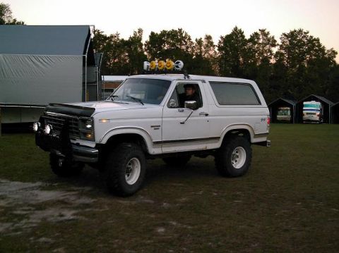 1986 Ford Bronco - A white Ford Bronco is parked on a grassy field with a driver inside.