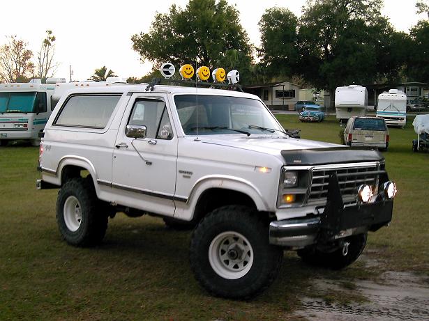 White Ford Bronco with lifted suspension, off-road tires, and a brush guard. Smiley face lights are mounted on the roof rack