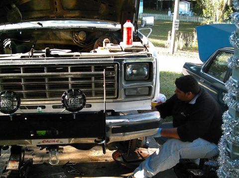 1986 Ford Bronco - A man works on the front bumper of a lifted Ford truck, which appears to be from the early 1980s, with another vehicle visible behind it.