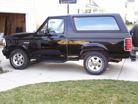 1996 Ford Bronco - Side profile of a black 1996 Ford Bronco with a spare tire on the back, parked in front of a garage.