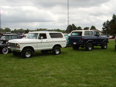 1978 Ford Bronco - Two Ford Broncos, a white one and a blue one, are parked on a grassy field under a cloudy sky.