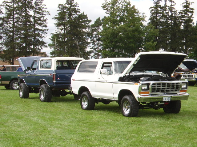 White 1978 Ford Bronco with hood open, parked next to a blue 1978 Ford Bronco. Another vehicle is visible behind them