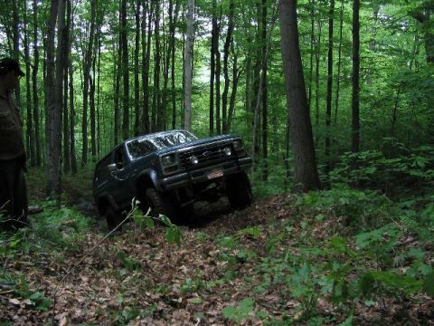 1986 Ford Bronco - A dark green SUV navigates a wooded trail, kicking up dirt as it goes.