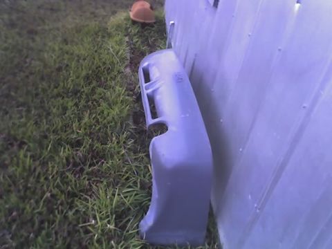 2000 Ford Escort - A gray car bumper is seen leaning against a light-colored wall next to a grassy area.