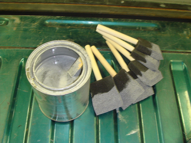 Can of gray paint with a stir stick and a group of foam brushes on a green ribbed surface