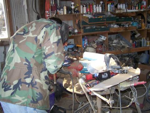 1995 Ford Bronco - A person wearing a welding mask and camouflage jacket works on a project in a garage filled with tools and automotive parts.