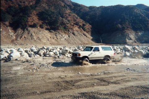 1988 Ford Bronco - White Ford Bronco navigates a water obstacle in a dry riverbed.