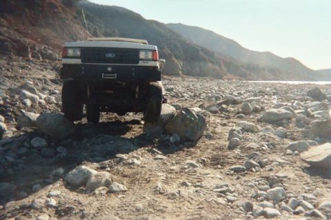 1988 Ford Bronco - A white Ford Bronco navigates a rocky, dry riverbed under a clear sky.