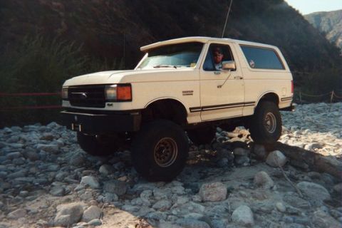 1988 Ford Bronco - A white Ford Bronco navigates a rocky terrain, with a driver visible behind the wheel.