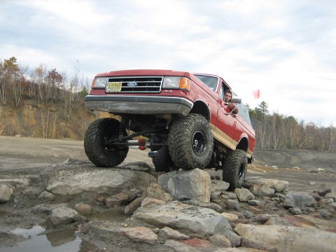 1990 Ford Bronco - A red Ford Bronco is shown off-roading over large rocks, demonstrating its lifted suspension and oversized tires.