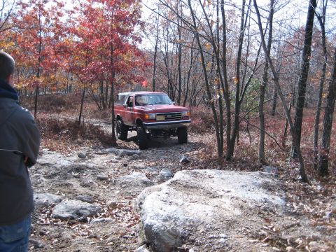 1990 Ford Bronco - A red SUV navigates a challenging rocky path through a wooded area during autumn.