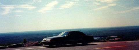 1997 Ford Crown Victoria - A black sedan is parked on the side of a road with a scenic view of water and trees.
