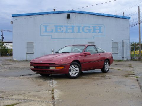 1991 Ford Probe - Low front three-quarter shot of a red Ford Probe hatchback coupe on cracked concrete, pop-up headlights down and silver multi-spoke alloys…