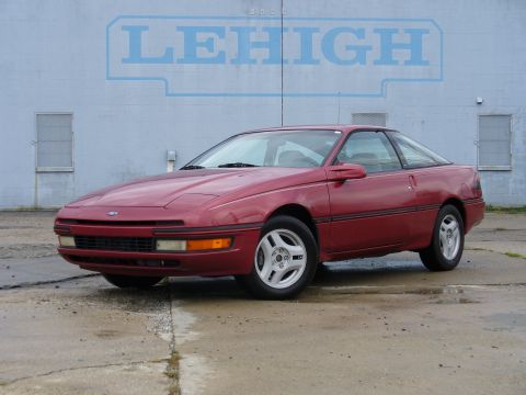 1991 Ford Probe - Clean red Ford Probe hatchback coupe photographed from the front three-quarter on a wet concrete lot in front of a pale block wall painted…