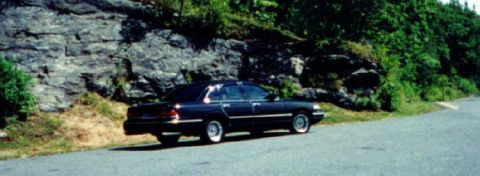 1997 Ford Crown Victoria - A dark 1997 Ford Crown Victoria sedan is parked on the side of a road with a rocky embankment and trees in the background.