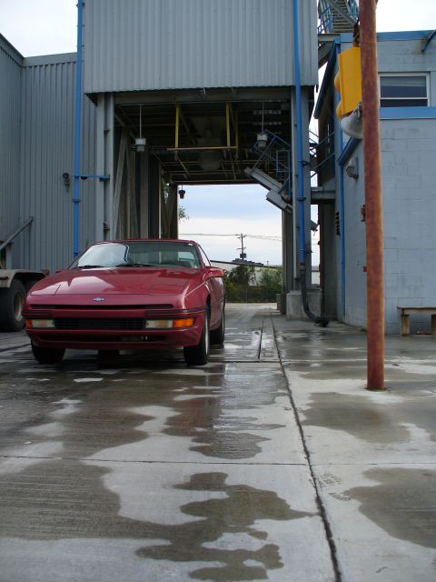 1991 Ford Probe - Low front-end view of a red first-generation Ford Probe parked on wet concrete in an industrial loading area, with the Ford oval on the smo…