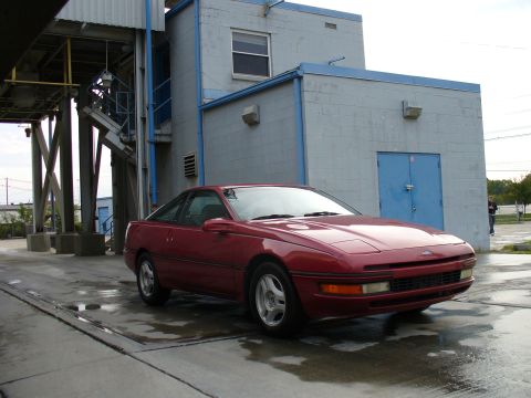 1991 Ford Probe - Red first-generation Ford Probe hatchback coupe photographed from a low 3/4 front angle on wet pavement beside a gray and blue cinder-block…