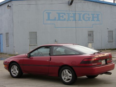 1991 Ford Probe - Red first-generation Ford Probe hatchback shot from a 3/4 rear angle beside a gray cinder-block wall painted with a large blue LEHIGH logo.…