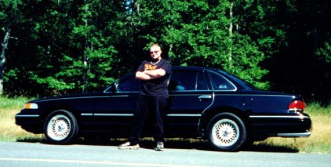 1997 Ford Crown Victoria - A man poses beside a dark-colored sedan parked on the side of a road with a wooded area behind it.