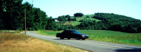 1997 Ford Crown Victoria - A black 1997 Ford Crown Victoria sedan travels down a winding country road under a clear blue sky.