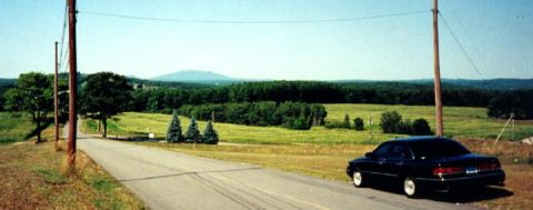 1997 Ford Crown Victoria - A black 1997 Ford Crown Victoria is seen from the rear as it travels down a country road with rolling hills in the background.