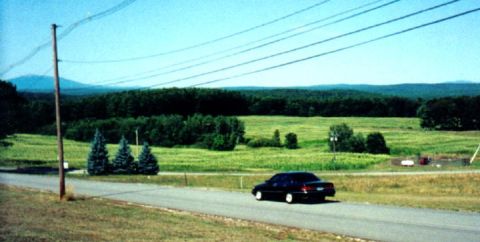 1997 Ford Crown Victoria - A dark sedan drives down a road in a rural landscape under a bright blue sky.