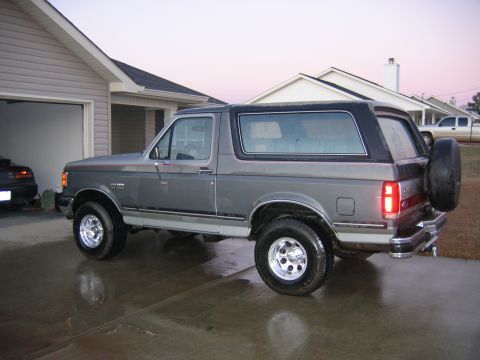 1989 Ford Bronco - A gray Ford Bronco with a black top is parked in a driveway next to a house.