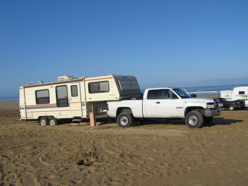 White 2001 Dodge 2500 pickup truck with a camper in the bed, towing a travel trailer on a sandy beach