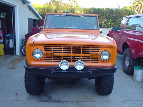 1977 Ford Bronco - Front view of an orange 1977 Ford Bronco, a custom bumper and auxiliary lights, parked next to a red truck.