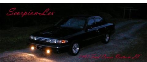 1997 Ford Crown Victoria - A black sedan is parked on a dirt road at night, illuminated by its headlights and fog lights.