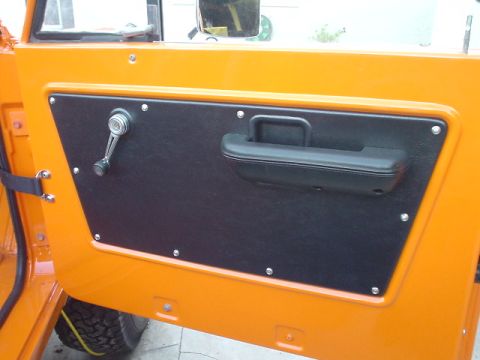1977 Ford Bronco - The interior door panel of an orange vehicle features a black armrest and a manual window crank.