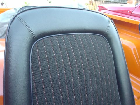 1977 Ford Bronco - Close-up of a black vinyl seat with red stitching, with parts of orange and red vehicles visible.