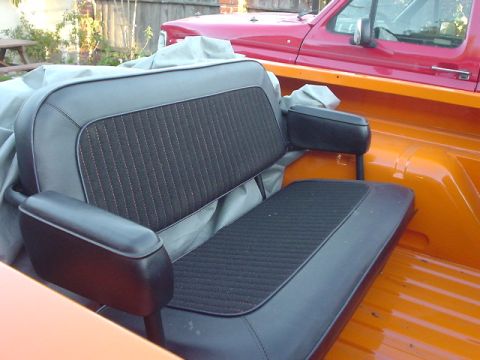 1977 Ford Bronco - Interior view of a rear bench seat with black upholstery, set against an orange interior and a red truck in the background.