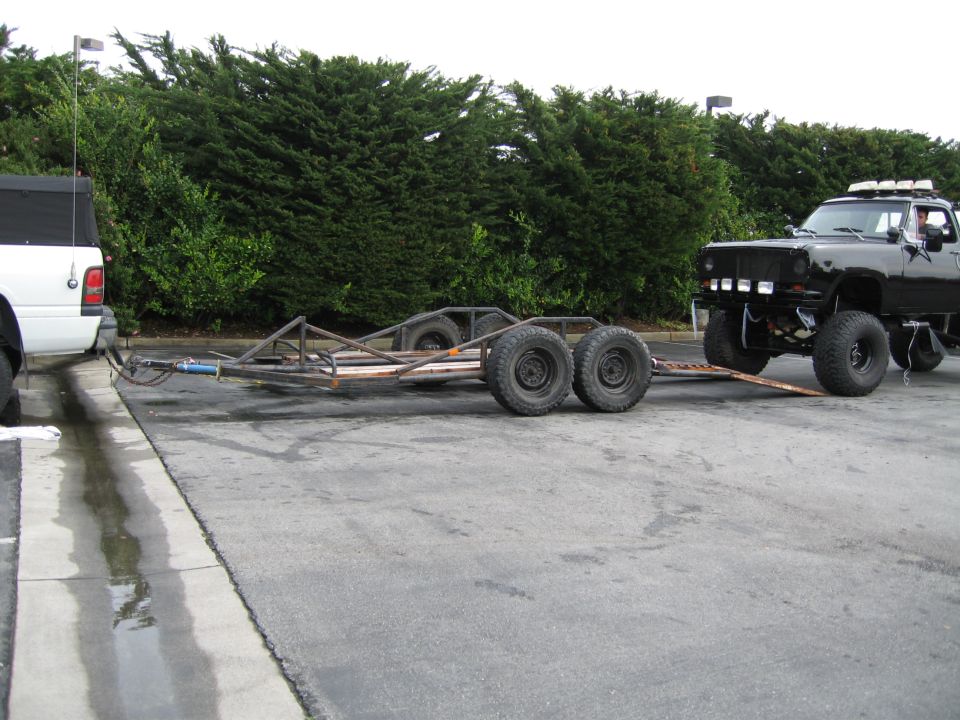 Black pickup truck with roof lights towing a custom trailer. White pickup truck is partially visible on the left