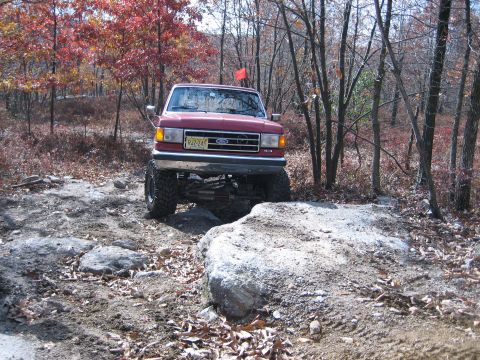 1990 Ford Bronco - Red 1990 Ford Bronco tackling an off-road course with large rocks and trees.