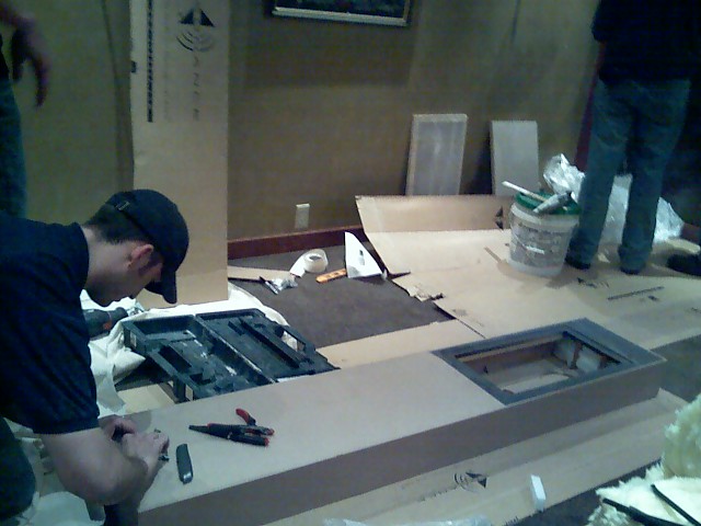 Construction scene with people working on a long, flat object on the floor, surrounded by cardboard boxes and tools