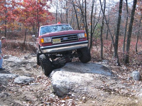 1990 Ford Bronco - A red 1990 Ford Bronco navigates a rocky obstacle during an off-road excursion in a wooded area.