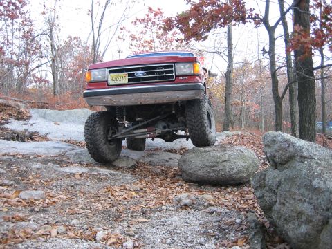 1990 Ford Bronco - A red Ford Bronco is shown off-roading over large rocks on a wooded trail.