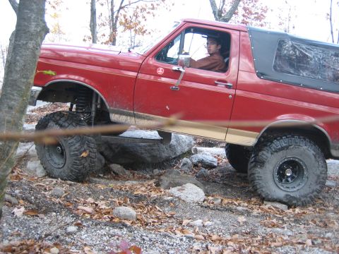 1990 Ford Bronco - A red 1990 Ford Bronco is shown off-roading over rocks and uneven ground.