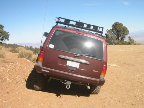 2000 Jeep Cherokee - A maroon 2000 Jeep Cherokee is angled on a dirt embankment under a clear blue sky, equipped with a roof rack and a 'NASA' sticker.