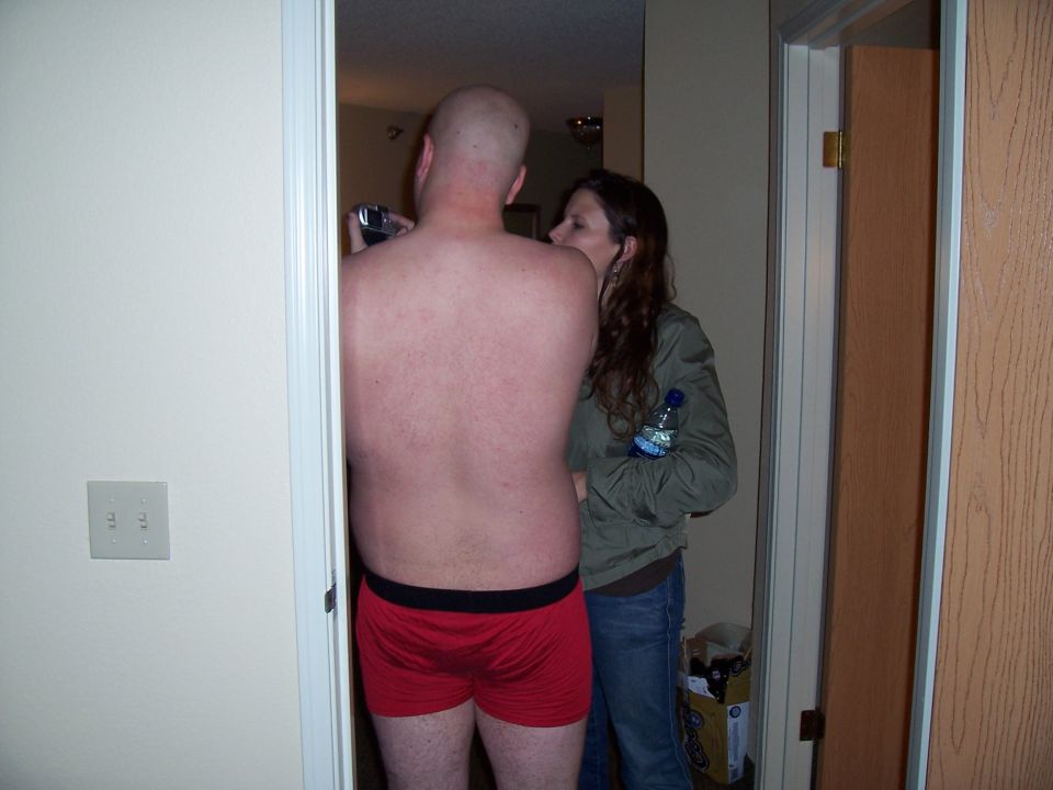 Bald man in red boxers holds camera, woman in green jacket holds water bottle, standing in doorway