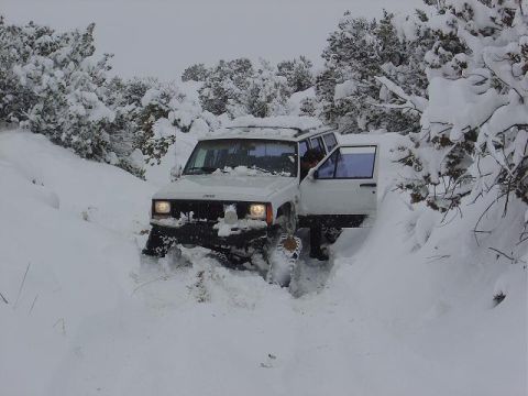 1986 Ford Bronco II - A white Jeep Cherokee XJ navigates through a snow-covered landscape with its driver's door ajar.